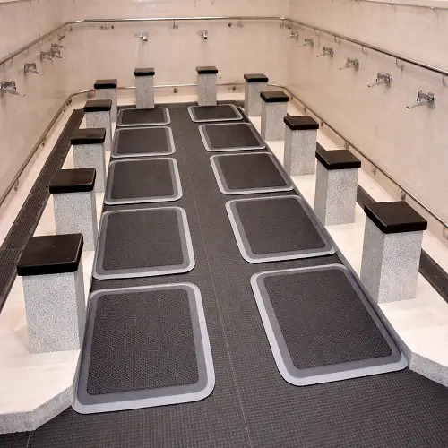 A clean mosque ablution area featuring multiple dark grey anti-slip ablution mats placed in front of washing stations and stone benches, designed to provide safe footing in wet conditions.