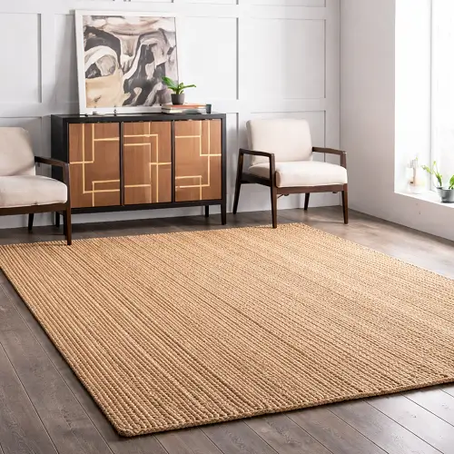 Modern living room featuring a large braided jute rug in natural beige tones, placed on wooden flooring and complemented by a stylish sideboard, armchairs, and minimalist décor for a clean and elegant interior look.