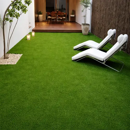 Outdoor backyard space covered with bright green nylon grass carpet, featuring two modern white lounge chairs and a wooden deck dining area in the background.