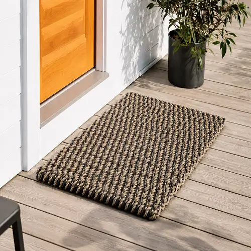 Durable outdoor mat with a textured woven pattern placed at a house entrance on a wooden deck, designed to remove dirt from shoes and provide a clean, slip-resistant surface outside the door.